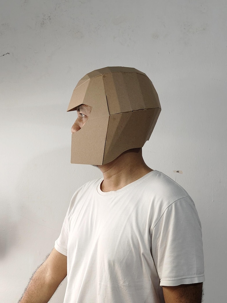 May include: A geometric helmet constructed from brown cardboard panels. The helmet features a faceted, angular design with a wide visor opening and a full-coverage shell that extends down to the jawline, showcasing a clean, modular construction.