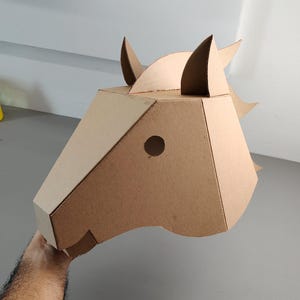 Puede incluir: Una m&aacute;scara geom&eacute;trica de cabeza de caballo construida con cart&oacute;n marr&oacute;n claro. La m&aacute;scara presenta un dise&ntilde;o angular y facetado con dos orejas erguidas, una abertura circular para el ojo y una crin estilizada en la parte posterior.