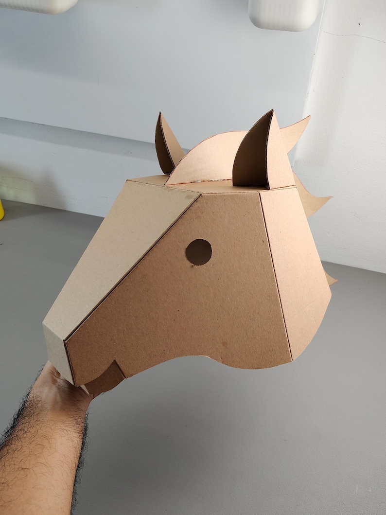 Puede incluir: Una m&aacute;scara geom&eacute;trica de cabeza de caballo construida con cart&oacute;n marr&oacute;n claro. La m&aacute;scara presenta un dise&ntilde;o angular y facetado con dos orejas erguidas, una abertura circular para el ojo y una crin estilizada en la parte posterior.