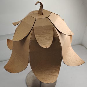 May include: A lamp shade constructed from brown corrugated cardboard, featuring a multi-layered petal design that flares outward. The structure is topped with a small, curved cardboard hook and is mounted on a circular, metallic silver base.