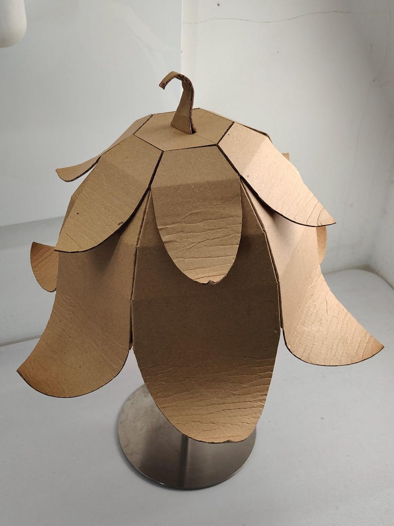 May include: A lamp shade constructed from brown corrugated cardboard, featuring a multi-layered petal design that flares outward. The structure is topped with a small, curved cardboard hook and is mounted on a circular, metallic silver base.