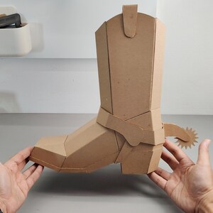 Puede incluir: Una escultura tridimensional de bota de vaquero construida con cart&oacute;n color canela. La bota presenta un dise&ntilde;o geom&eacute;trico y facetado con una espuela prominente unida al &aacute;rea del tal&oacute;n. La estructura se compone de m&uacute;ltiples paneles de cart&oacute;n planos.