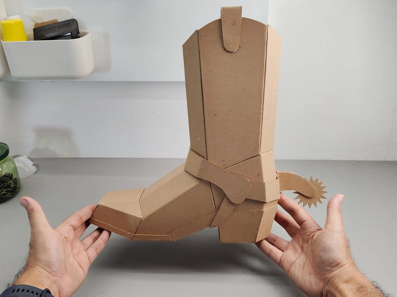 Puede incluir: Una escultura tridimensional de bota de vaquero construida con cart&oacute;n color canela. La bota presenta un dise&ntilde;o geom&eacute;trico y facetado con una espuela prominente unida al &aacute;rea del tal&oacute;n. La estructura se compone de m&uacute;ltiples paneles de cart&oacute;n planos.