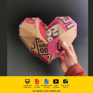 May include: A 3D heart sculpture constructed from recycled cardboard packaging with pink and brown printed text and graphics. The geometric, faceted object features visible seams and folds. Below the heart, text reads 3D HEART LOVE TEMPLATE.