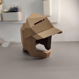 May include: A brown cardboard helmet with a visor and angular design. The helmet has a rectangular opening for the eyes and a chin guard. The helmet is made of folded cardboard and sits on a gray surface.