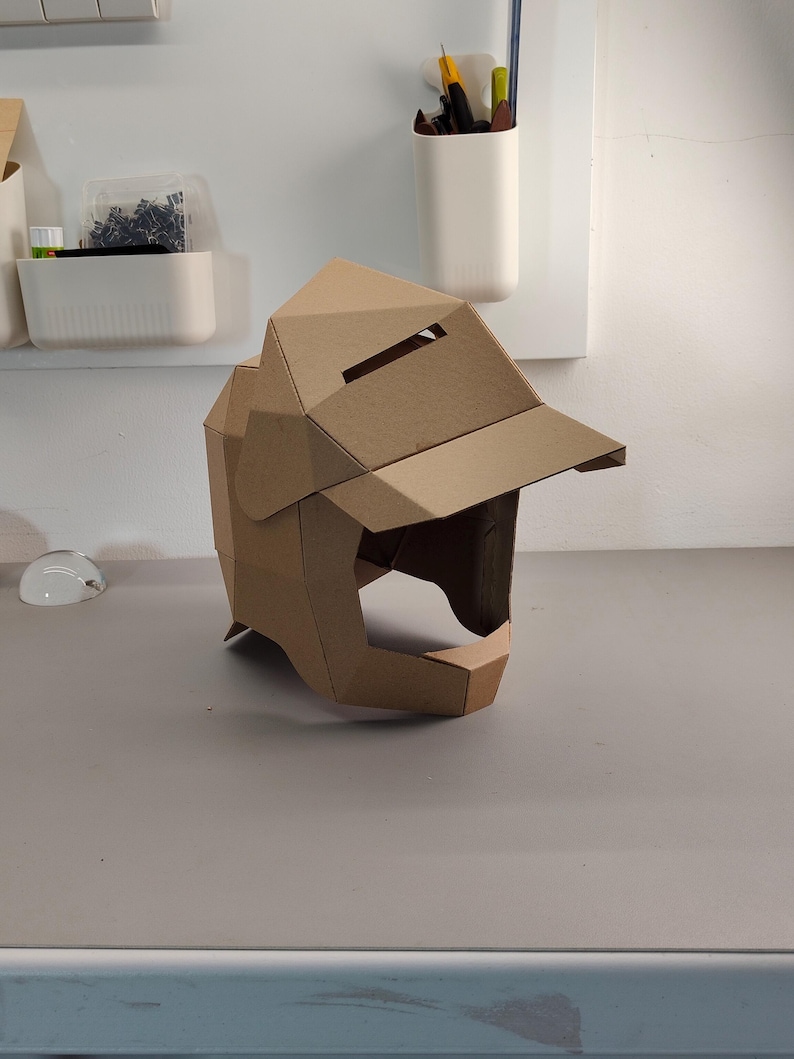 May include: A brown cardboard helmet with a visor and angular design. The helmet has a rectangular opening for the eyes and a chin guard. The helmet is made of folded cardboard and sits on a gray surface.