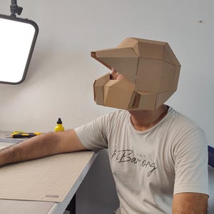 Op de afbeelding: Een geometrische helm gemaakt van bruine kartonnen panelen, met een vizierachtige uitstulping en een hoekig, gefacetteerd ontwerp. De helm wordt op het hoofd gedragen en vertoont een gestructureerd uiterlijk met meerdere panelen en naden.