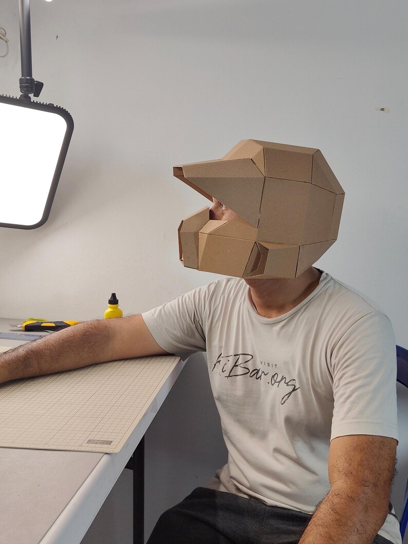 Op de afbeelding: Een geometrische helm gemaakt van bruine kartonnen panelen, met een vizierachtige uitstulping en een hoekig, gefacetteerd ontwerp. De helm wordt op het hoofd gedragen en vertoont een gestructureerd uiterlijk met meerdere panelen en naden.