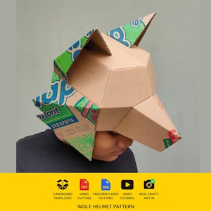 May include: A geometric wolf helmet constructed from brown cardboard with green and blue printed packaging details on the ears and sides. The helmet features a pointed snout and upright ears, showcasing a faceted, angular design made from recycled materials.