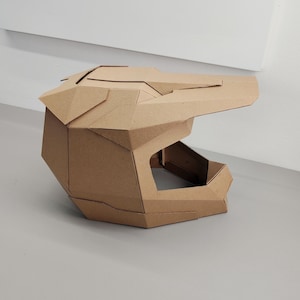 May include: A geometric helmet constructed from brown corrugated cardboard. The helmet features a prominent visor, a wide face opening, and angular, faceted panels that form a structured, multi-piece design with visible seams throughout.