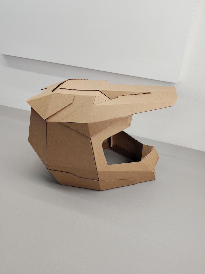 May include: A geometric helmet constructed from brown corrugated cardboard. The helmet features a prominent visor, a wide face opening, and angular, faceted panels that form a structured, multi-piece design with visible seams throughout.