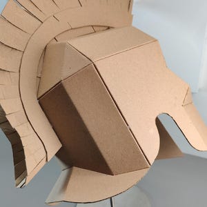 May include: A tan cardboard helmet constructed with geometric panels and a tall, fringed crest that arches over the top. The helmet features a rigid, angular design with a wide face opening and a flared neck guard, crafted from plain brown paperboard.