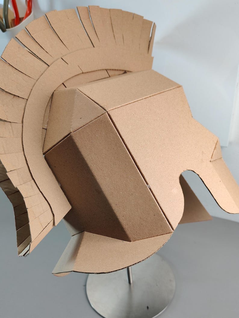 May include: A tan cardboard helmet constructed with geometric panels and a tall, fringed crest that arches over the top. The helmet features a rigid, angular design with a wide face opening and a flared neck guard, crafted from plain brown paperboard.