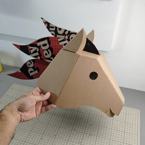 Puede incluir: Una escultura de cabeza de caballo en tres dimensiones construida con cart&oacute;n color canela. La crin est&aacute; hecha de piezas de cart&oacute;n superpuestas con patrones en negro, rojo y blanco, con el texto visible "pelly" y "ed" en una fuente negrita.