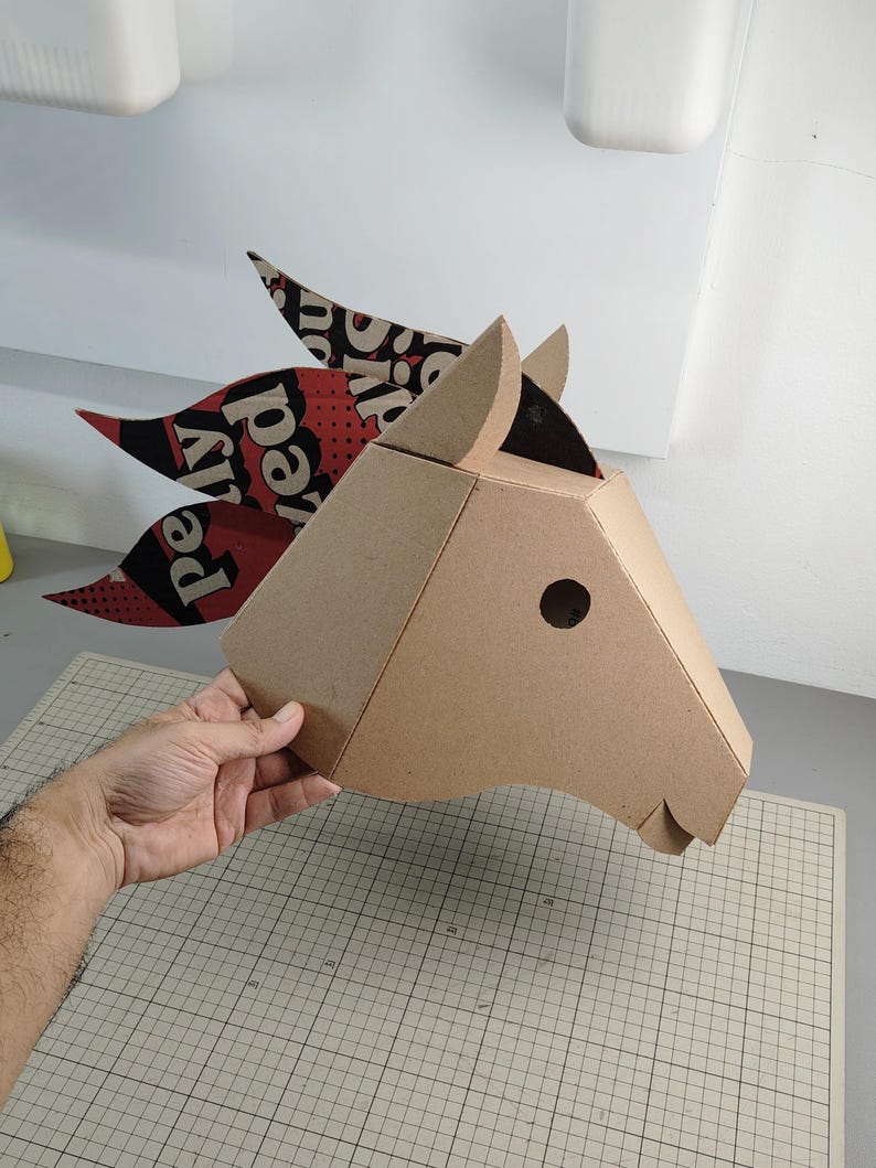Puede incluir: Una escultura de cabeza de caballo en tres dimensiones construida con cart&oacute;n color canela. La crin est&aacute; hecha de piezas de cart&oacute;n superpuestas con patrones en negro, rojo y blanco, con el texto visible "pelly" y "ed" en una fuente negrita.