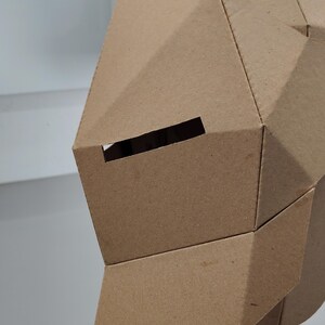 May include: A geometric cardboard mask with a rectangular eye opening. The mask is constructed from folded, brown cardboard pieces, creating a faceted surface. The design suggests a craft project or costume element.