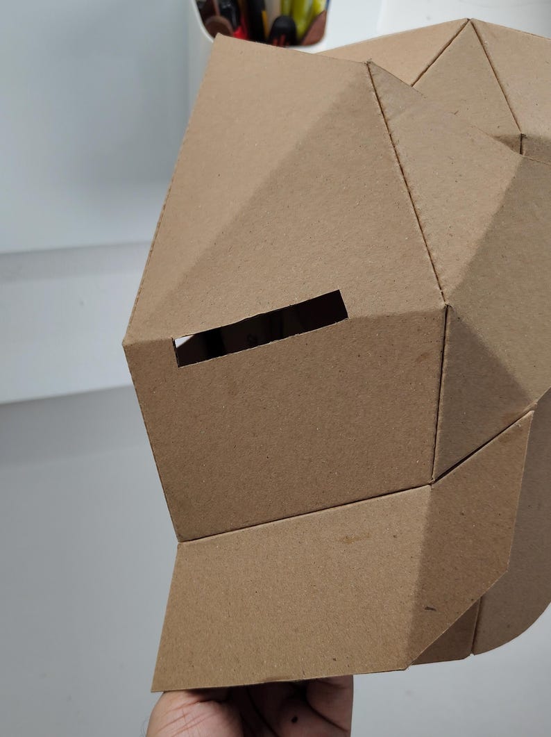 May include: A geometric cardboard mask with a rectangular eye opening. The mask is constructed from folded, brown cardboard pieces, creating a faceted surface. The design suggests a craft project or costume element.