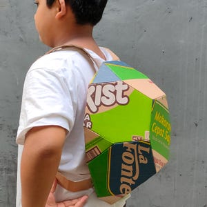 May include: A child wearing a white shirt is wearing a backpack made from cardboard boxes. The backpack is decorated with various food product labels, including "Kist" and "La Fonte".