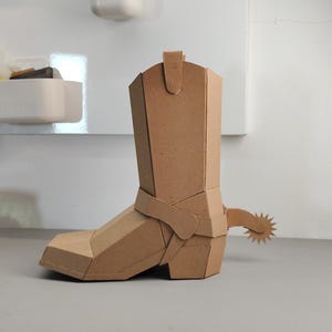 Puede incluir: Una escultura tridimensional de bota de vaquero construida con cart&oacute;n marr&oacute;n. La bota presenta un dise&ntilde;o geom&eacute;trico y facetado con una espuela prominente unida al tal&oacute;n. La estructura est&aacute; compuesta por paneles planos y angulares unidos.
