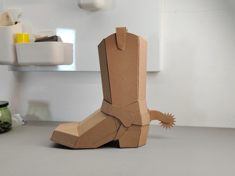 Puede incluir: Una escultura tridimensional de bota de vaquero construida con cart&oacute;n marr&oacute;n. La bota presenta un dise&ntilde;o geom&eacute;trico y facetado con una espuela prominente unida al tal&oacute;n. La estructura est&aacute; compuesta por paneles planos y angulares unidos.