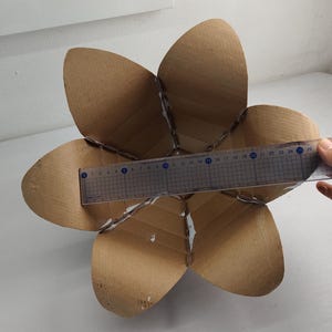 May include: A brown cardboard structure shaped like a six-petaled flower. A clear plastic ruler is placed across the center, showing a width of approximately 9.5 inches from the edge of one petal to the opposite side, with visible adhesive seams.