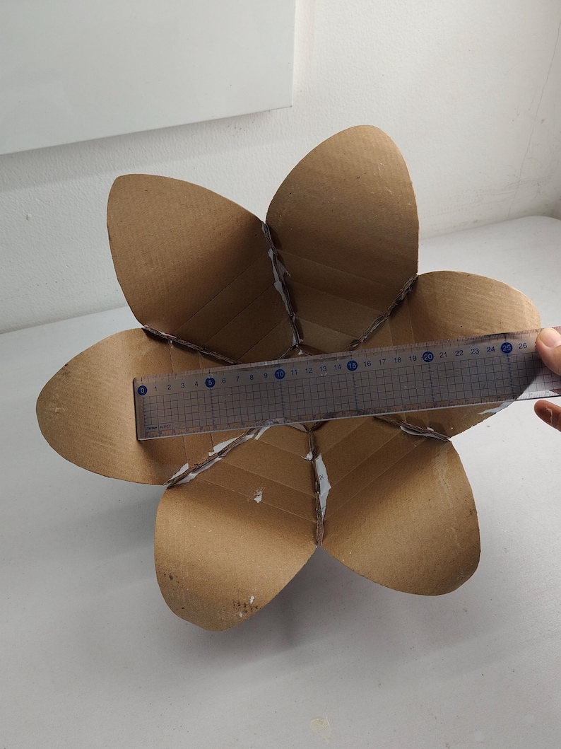 May include: A brown cardboard structure shaped like a six-petaled flower. A clear plastic ruler is placed across the center, showing a width of approximately 9.5 inches from the edge of one petal to the opposite side, with visible adhesive seams.
