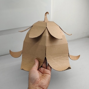 May include: A light brown cardboard structure shaped like a bell-shaped flower with multiple layered petals. The object features a central stem-like protrusion at the top and curved, outward-pointing petal edges, constructed from flat, tan-colored material.