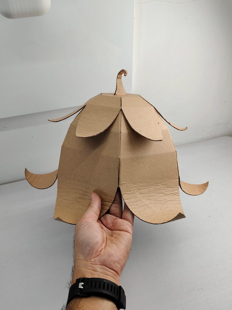 May include: A light brown cardboard structure shaped like a bell-shaped flower with multiple layered petals. The object features a central stem-like protrusion at the top and curved, outward-pointing petal edges, constructed from flat, tan-colored material.