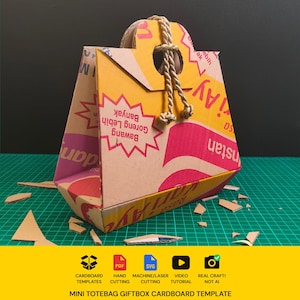 Puede incluir: Una mini bolsa de regalo tipo tote hecha de cartón reciclado con texto impreso en amarillo, rosa y rojo. La caja tiene forma triangular, un asa troquelada y un asa de cuerda beige gruesa y trenzada. Hay restos de cartón cerca.