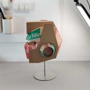 May include: A sculptural helmet constructed from brown cardboard packaging featuring visible green and red printed text including the word Mie. The structure is mounted on a silver metal stand with a circular base and features a circular cutout.