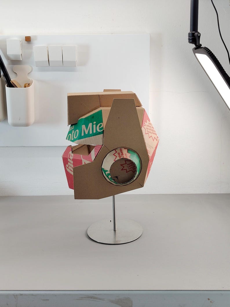 May include: A sculptural helmet constructed from brown cardboard packaging featuring visible green and red printed text including the word Mie. The structure is mounted on a silver metal stand with a circular base and features a circular cutout.