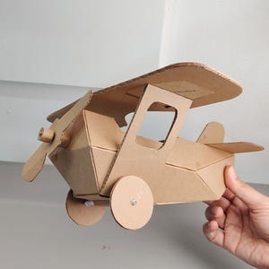May include: A biplane model constructed from brown corrugated cardboard. The aircraft features a top wing, a front propeller, two circular side wheels, and a tail fin. The structure is assembled with visible tabs and slots, showcasing a geometric design.