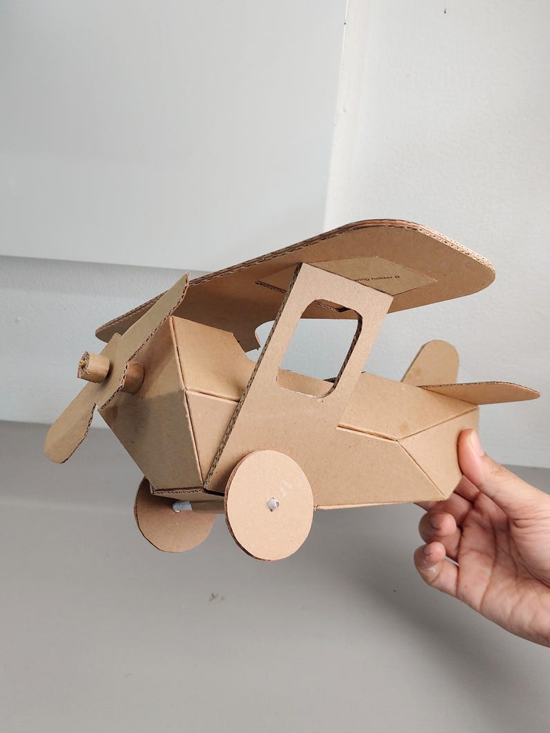 May include: A biplane model constructed from brown corrugated cardboard. The aircraft features a top wing, a front propeller, two circular side wheels, and a tail fin. The structure is assembled with visible tabs and slots, showcasing a geometric design.