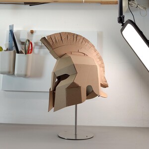 May include: A tan cardboard Spartan-style helmet featuring a prominent crest, angular face guard, and eye openings. The helmet is mounted on a silver metal stand with a circular base, showcasing a geometric construction made from layered cardboard panels.