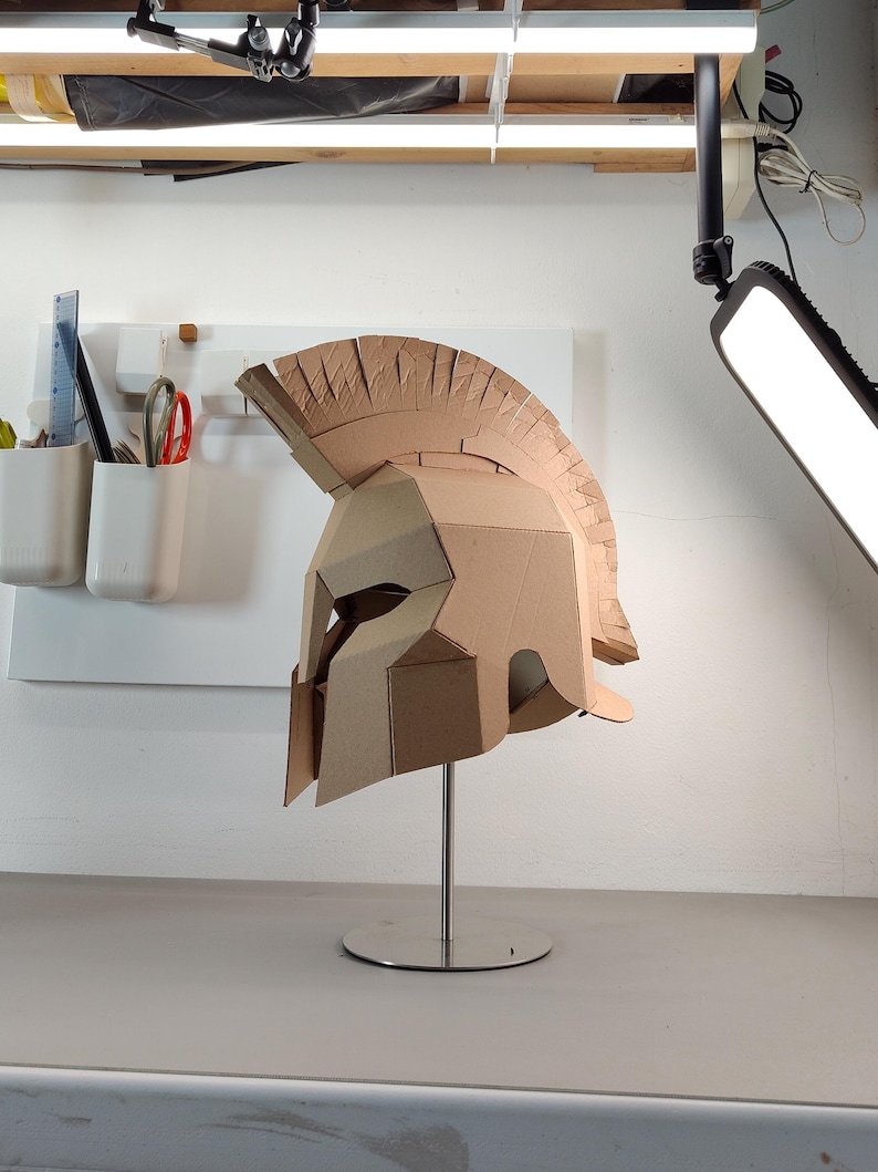 May include: A tan cardboard Spartan-style helmet featuring a prominent crest, angular face guard, and eye openings. The helmet is mounted on a silver metal stand with a circular base, showcasing a geometric construction made from layered cardboard panels.