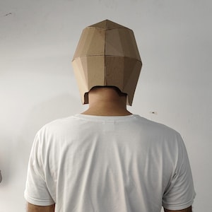 May include: A geometric, faceted mask constructed from light brown cardboard, featuring a multi-panel design that covers the upper head and sides. The mask is worn over a plain white short-sleeved t-shirt, showcasing a simple, angular structure.