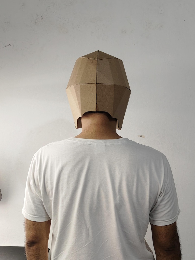 May include: A geometric, faceted mask constructed from light brown cardboard, featuring a multi-panel design that covers the upper head and sides. The mask is worn over a plain white short-sleeved t-shirt, showcasing a simple, angular structure.