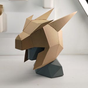 Könnte beinhalten: Eine geometrische Drachenkopfskulptur aus Pappe mit scharfen Winkeln und Spitzen. Der Drachenkopf ist beige und sitzt auf einem grauen geometrischen Sockel. Die Skulptur ist ein kreatives Bastelprojekt.