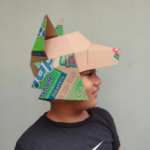 May include: A handmade cardboard wolf mask with green and blue accents. The mask is constructed from recycled cardboard boxes, featuring the text "Instant" and other Indonesian text. The wolf's head is tan with pointed ears.