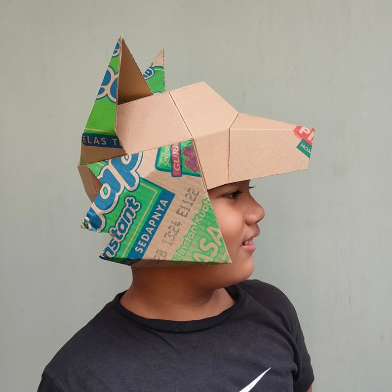 May include: A handmade cardboard wolf mask with green and blue accents. The mask is constructed from recycled cardboard boxes, featuring the text "Instant" and other Indonesian text. The wolf's head is tan with pointed ears.