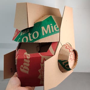 May include: A geometric mask constructed from repurposed brown cardboard packaging. The structure features angular panels with visible red and green printed text including 'Rasa Soto Mie' and 'Saus Cabe'. A circular cardboard protrusion extends from the side.