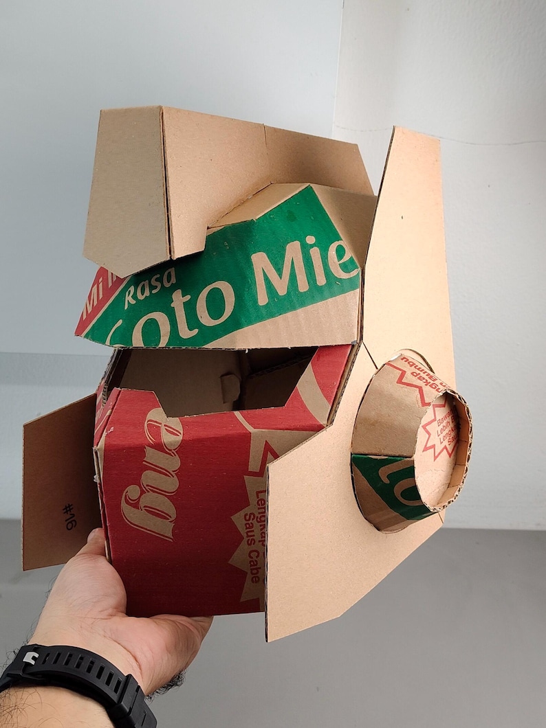 May include: A geometric mask constructed from repurposed brown cardboard packaging. The structure features angular panels with visible red and green printed text including 'Rasa Soto Mie' and 'Saus Cabe'. A circular cardboard protrusion extends from the side.