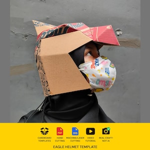 May include: A geometric eagle helmet constructed from brown cardboard with printed red and black graphic patterns. The helmet features a pointed beak and angular head structure. Below the helmet, a white face mask displays colorful graphic text.