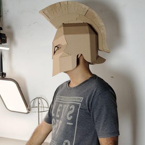 May include: A tan cardboard Spartan-style helmet featuring a prominent crest and angular face guard. The helmet is constructed from layered corrugated material with visible seams and edges, designed to cover the head and upper face area.