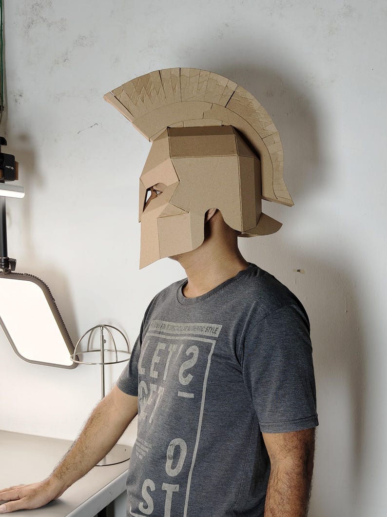 May include: A tan cardboard Spartan-style helmet featuring a prominent crest and angular face guard. The helmet is constructed from layered corrugated material with visible seams and edges, designed to cover the head and upper face area.