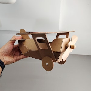 May include: A model aeroplane constructed from brown corrugated cardboard. The aircraft features a main wing, a propeller attached to the nose, a tail section, and two circular wheels on the sides. The structure is assembled with visible edges.