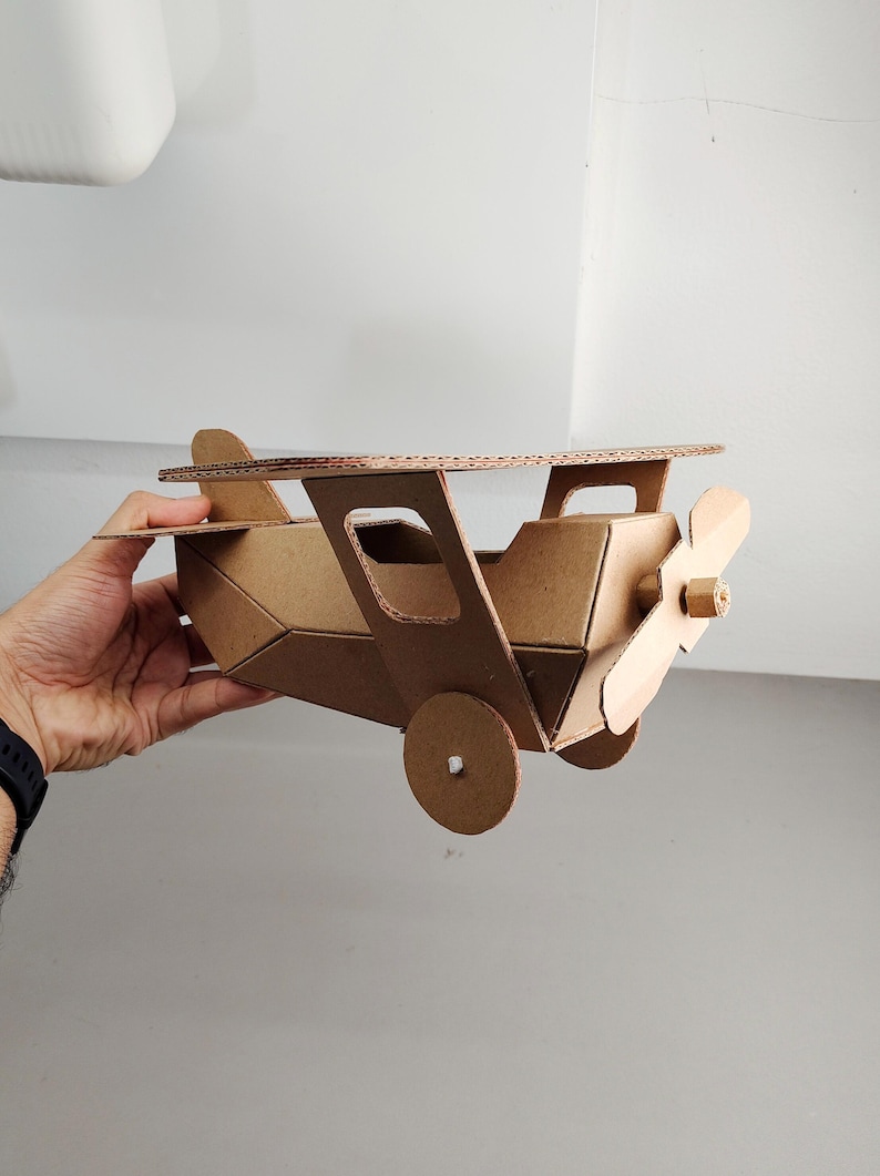 May include: A model aeroplane constructed from brown corrugated cardboard. The aircraft features a main wing, a propeller attached to the nose, a tail section, and two circular wheels on the sides. The structure is assembled with visible edges.