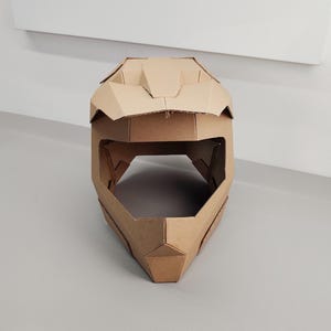 May include: A full-face helmet constructed from brown corrugated cardboard. The helmet features a geometric, angular design with a prominent visor, a wide central eye opening, and a pointed chin guard, all assembled from flat cardboard panels.
