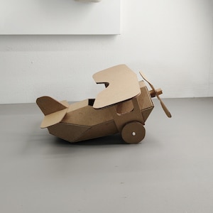 May include: A brown cardboard biplane model featuring two stacked wings, a front-mounted propeller, and a tail assembly. The structure is constructed from folded corrugated cardboard panels with circular wheels attached to the sides of the fuselage.