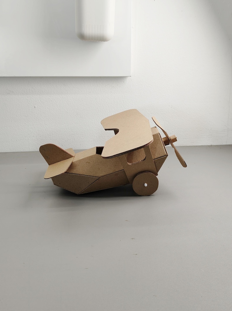 May include: A brown cardboard biplane model featuring two stacked wings, a front-mounted propeller, and a tail assembly. The structure is constructed from folded corrugated cardboard panels with circular wheels attached to the sides of the fuselage.